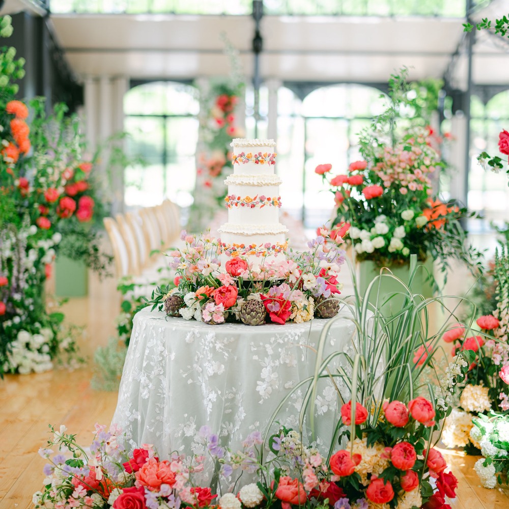 Spitzen-Overlay-Tischdecke in Hellgrau mit weißer Kirschblütenstickerei auf rundem Kuchentisch, vierstöckige Hochzeitstorte mit bunten Blumenkränzen, umgeben von Korallen- und Pinkblumen, gläsernes Gewächshaus