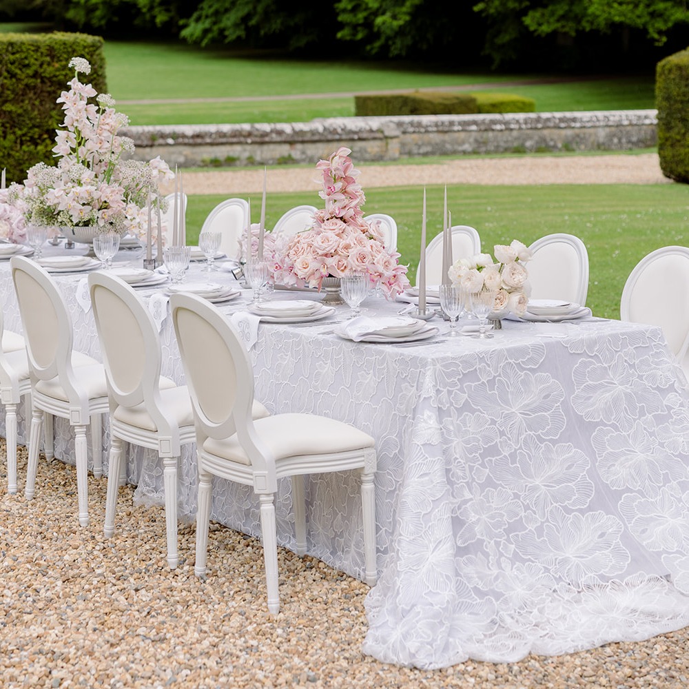 Luxuriöse Spitzen-Overlay-Tischdecke in Hellgrau mit weißer Ginkgoblatt-Stickerei auf einem langen Hochzeitstisch im Freien, altrosa Rosen- und Orchideengestecke, Louis-XVI-Stühle, Schlossgarten
