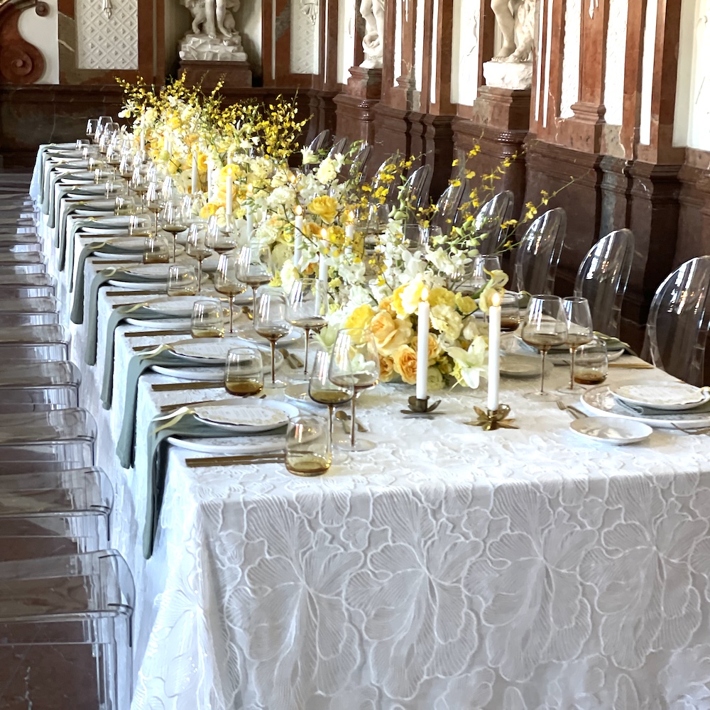 Langer Banketttisch mit weißer Ginkgoblatt-Spitzen-Overlay-Tischdecke, gelbe Rosen- und Oncidium-Orchideengestecke, salbeigrüne Leinenservietten, Ghost-Chairs, Barockpalais mit Marmorstatuen