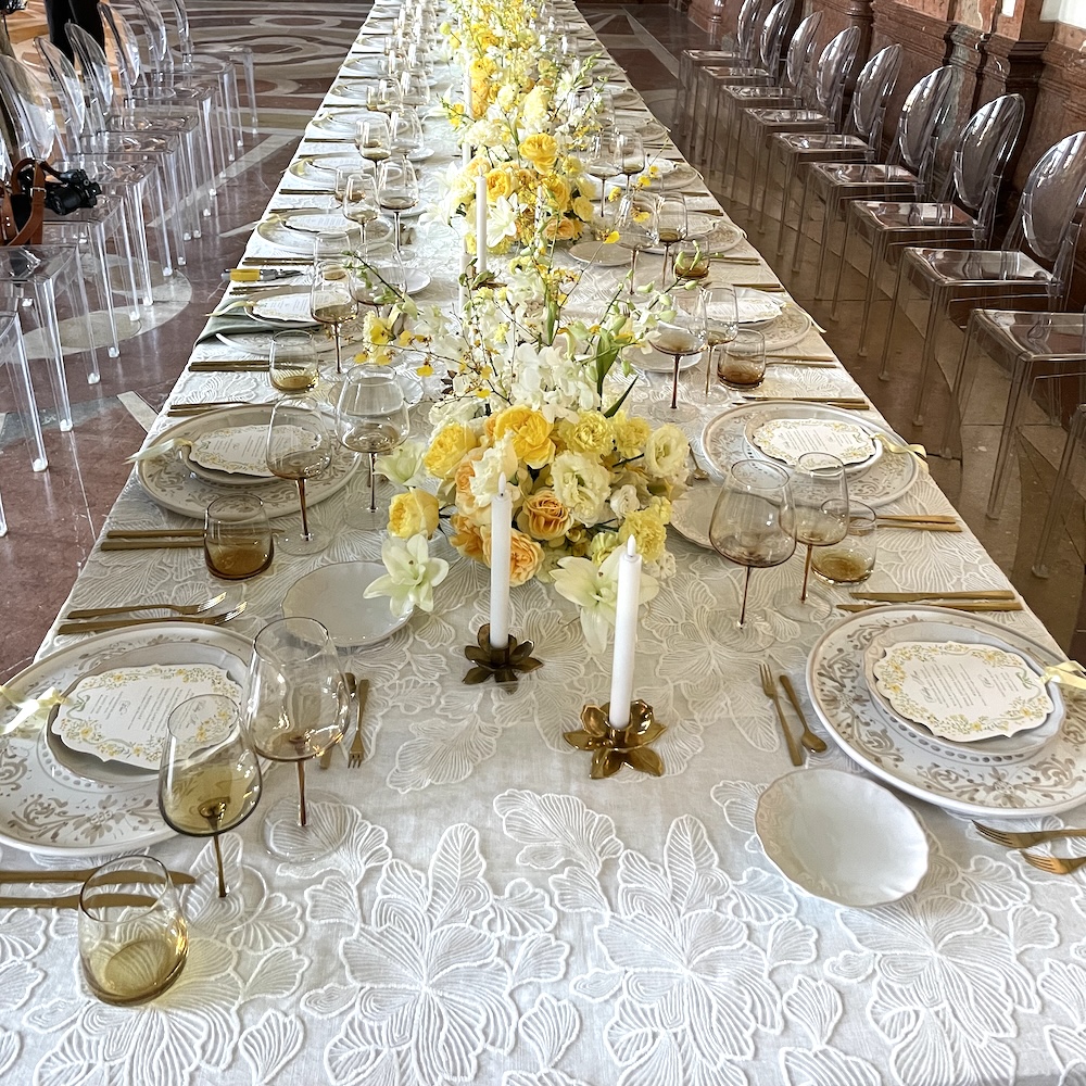 Langer Festtisch mit weißer Ginkgoblatt-Spitzen-Overlay-Tischdecke von oben, gelbe Rosen- und Orchideengestecke als Blumenläufer, goldenes Besteck und bernsteinfarbene Gläser, Marmorsaal Barockpalais